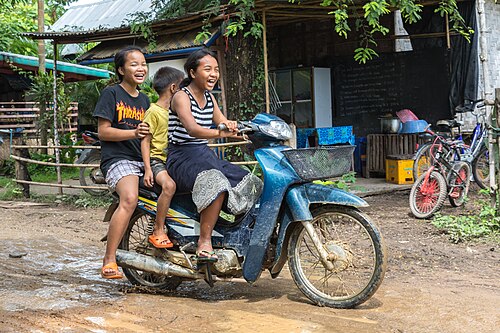 Girl riding a motorcycle with two passengers laughing on the muddy slope of Don Khon